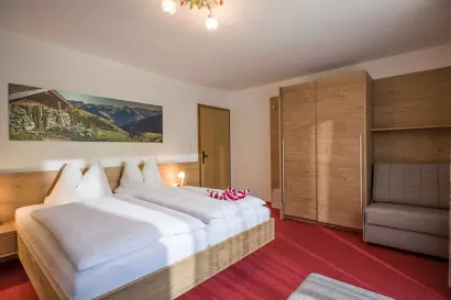 Et lyst hotelværelse med dobbeltseng, rødt tæppe, træklædeskab, lille grå sofa og et landskabsfoto på væggen. Sollys strømmer ind gennem et skjult vindue og skaber en varm og indbydende atmosfære.