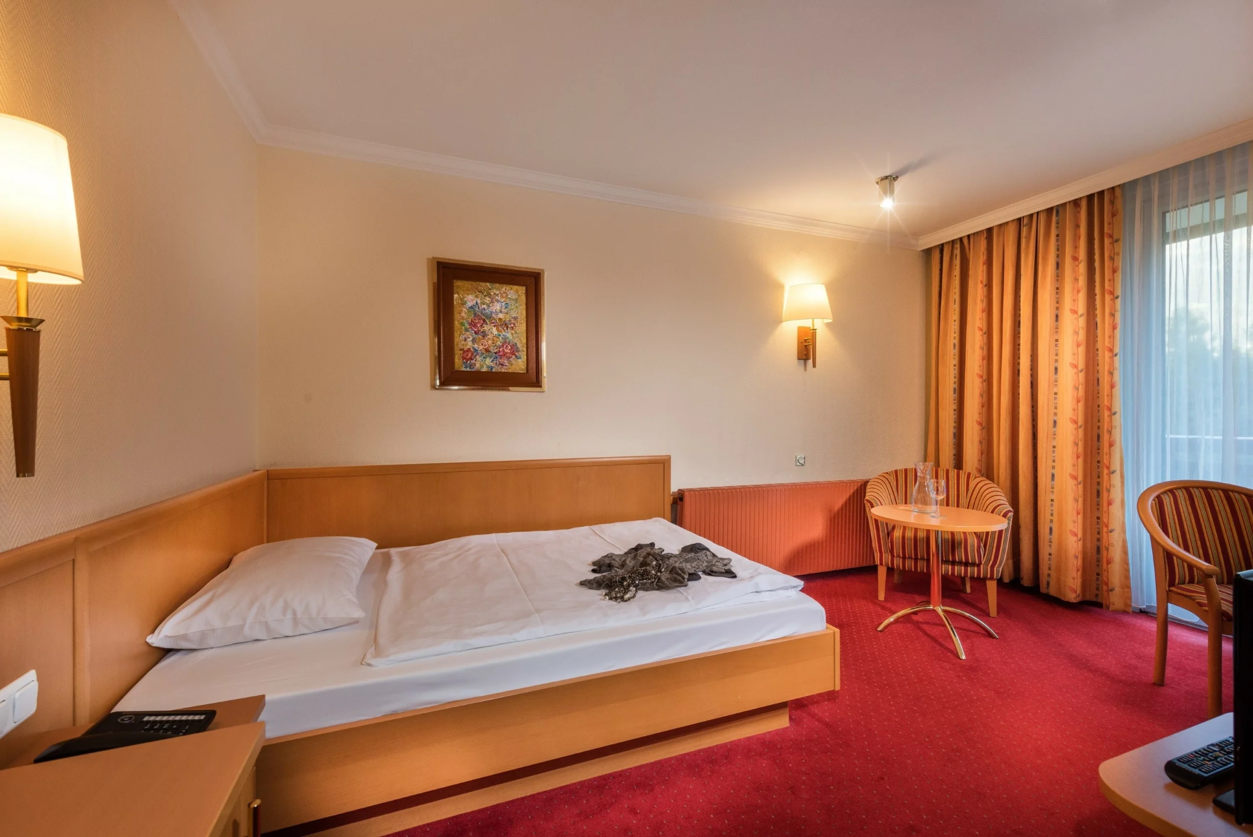 En pænt redt seng på et hotelværelse med rødt tæppe, træmøbler, et lille rundt bord med to stole, væglamper, et indrammet maleri, fortrukne gardiner og en skydedør af glas, der lader naturligt lys komme ind.