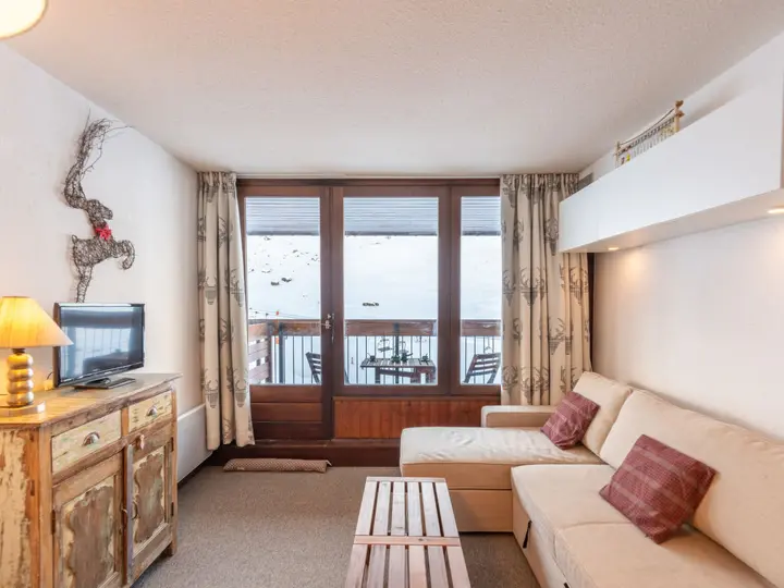 En hyggelig stue med en beige sofa, røde hynder, rustikke træmøbler og et tv. Store glasdøre åbner ud til en balkon med udendørs siddepladser med udsigt over et snedækket bjerglandskab.
