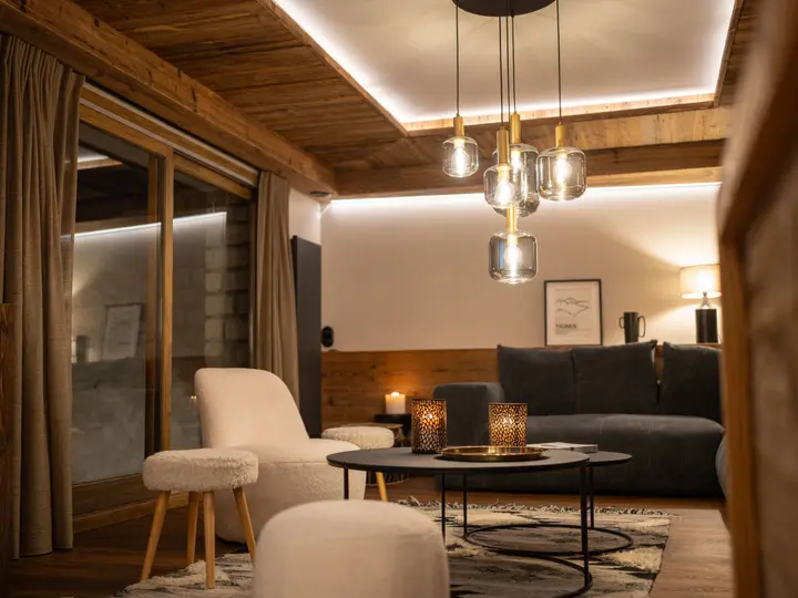 En hyggelig stue med træloft og -vægge, moderne pendellamper, en mørk sofa, lyse lænestole, runde sofaborde med dekorative bakker og lysestager samt store vinduer med gardiner.