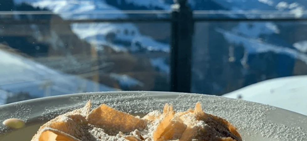 En skive dessert, eventuelt et wienerbrød eller kage, drysset med flormelis på en tallerken. I baggrunden er de snedækkede bjerge i Saalbach synlige under en klar blå himmel, så det føles som et perfekt skiferie øjeblik.