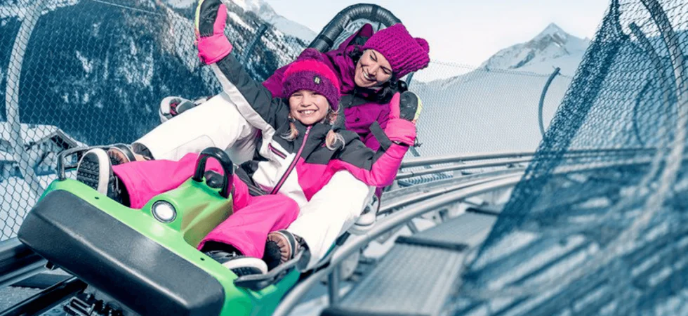 To mennesker bundtet i vintertøj kører på en grøn alpine coaster, smilende og rækker hænderne op. Sneklædte bjerge og et sikkerhedsnet hegn er synlige mod den klare blå himmel i Westendorf, og fanger glæden hos skirejser selv off-piste.
