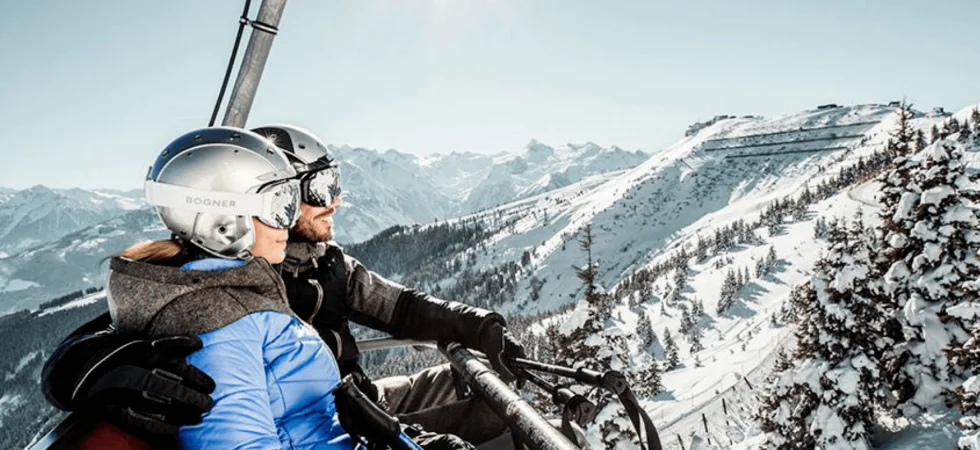To personer iført skihjelme og vinterudstyr sidder på en skilift i Wagrain og stirrer på snedækkede bjerge under en strålende sol. Landskabet er oversået med træer under en klar blå himmel, der fanger den fredfyldte skønhed i de alpine omgivelser.