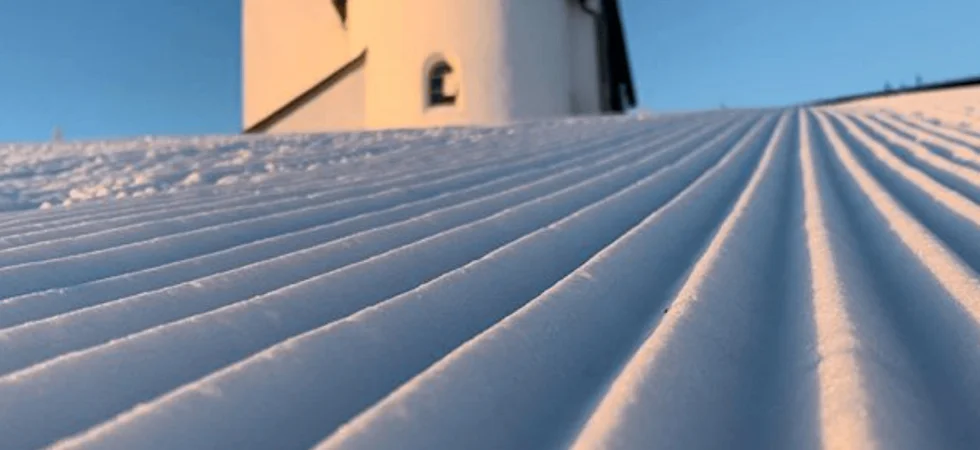 En snedækket skråning med pæne, parallelle riller fører op til en hvid bygning med et lille tårn, sat mod en klar blå himmel - en perfekt scene for din næste skiferie i Westendorf.