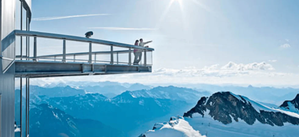 To personer står på en udsigtsplatform, der strækker sig fra en snedækket bygning i Saalbach, med udsigt over en panoramisk bjergkæde under en klar blå himmel og sol. Snedækkede tinder og en stor himmel skaber den fredfyldte, betagende udsigt, der er typisk for skirejseoplevelser.