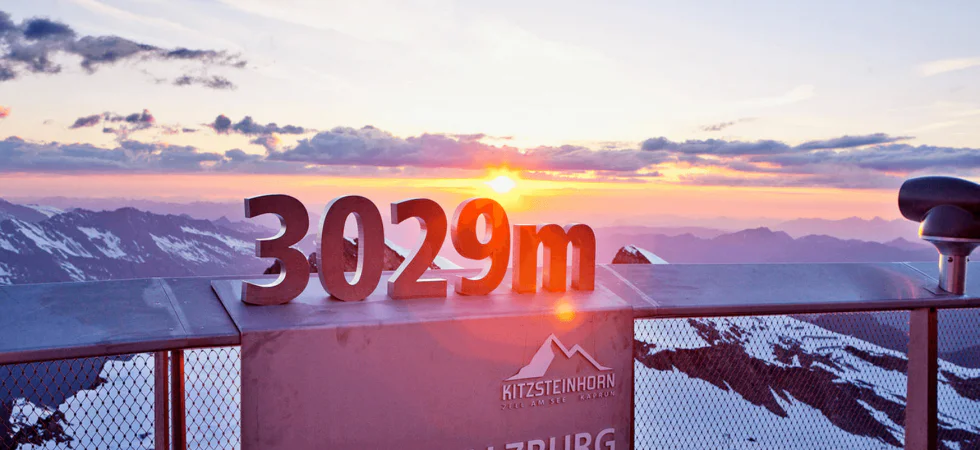 Oplev den betagende udsigt over solopgangen fra Kitzsteinhorn-gletsjeren på toppen af Salzburg, Østrig. På 3029m er snedækkede bjerge og skyer badet i solens varme skær – en perfekt start på dit skiferieeventyr, kun en kort rejse fra Wagrain.