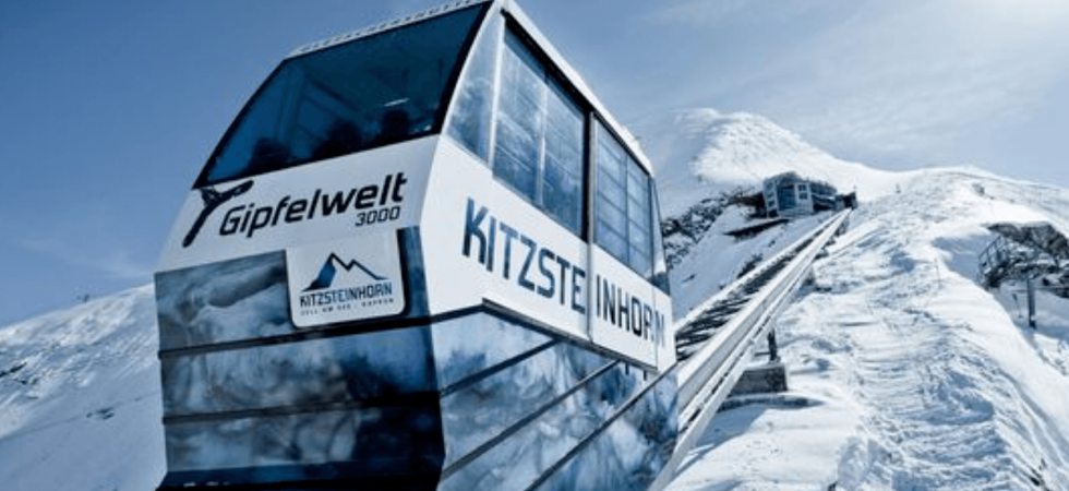 En svævebane mærket Kitzsteinhorn Gipfelwelt 3000 bestiger et snedækket bjerg under en klar blå himmel, der minder om de maleriske landskaber, man kan finde på skirejsedestinationer som Westendorf. Snedækkede tinder og en bjergstation pryder baggrunden med et gult retningsskilt ved siden af banen.