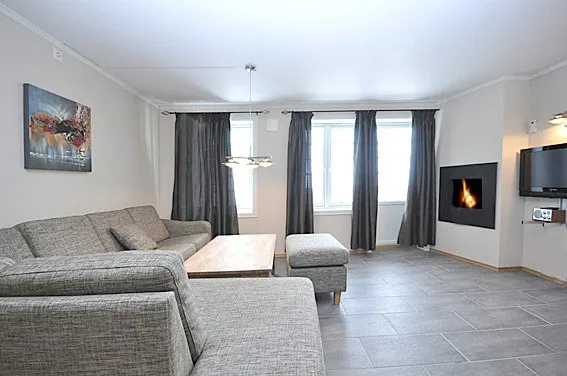 En moderne stue, der minder om en smart Wagrain skiferie, med en grå sektionssofa, sofabord i træ og matchende skammel. Store vinduer kan prale af grå gardiner, mens et vægmonteret tv og pejs pryder den ene side, og et moderne maleri fuldender den hyggelige skihytte-atmosfære.