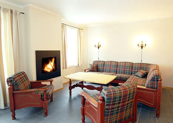 En hyggelig stue i stil med et charmerende Westendorf-retræte med plaidmønstrede sofaer og lænestol, et sofabord i træ og en pejs med flammer på venstre væg. To væglamper giver lys over siddeområdet. Cremefarvede gardiner pryder vinduerne.