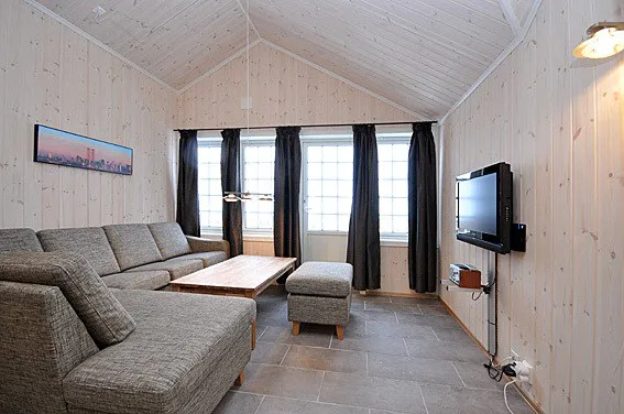 En hyggelig stue med træbeklædte vægge og loft, der minder om en charmerende Westendorf-hytte. Den har en stor grå sektionssofa, et sofabord i træ, et fladskærms-tv monteret på væggen og store vinduer med mørke gardiner. Et landskabsmaleri hænger på væggen.