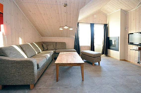En hyggelig stue med skrå trælofter og vægge, der minder om en Westendorf skiferie. Den har en grå L-formet sofa, et sofabord i træ, en matchende skammel, et vægmonteret tv, en pejs og store vinduer prydet med sorte gardiner. Gulvet er elegant flisebelagt.
