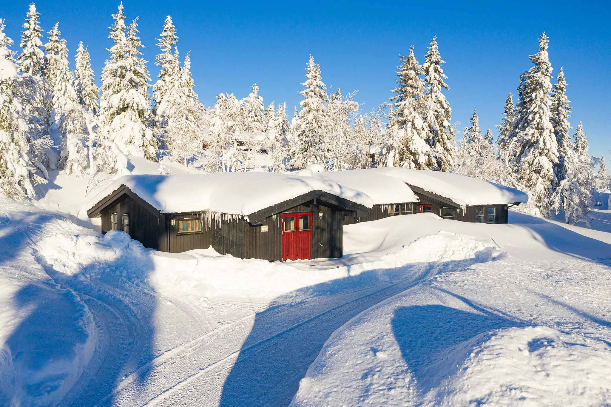 En hyggelig hytte med røde døre ligger i det snedækkede landskab i Westendorf, omgivet af snefyldte træer under en klar blå himmel. Dækspor er synlige i sneen, hvilket fører til denne perfekte skiferieferie.