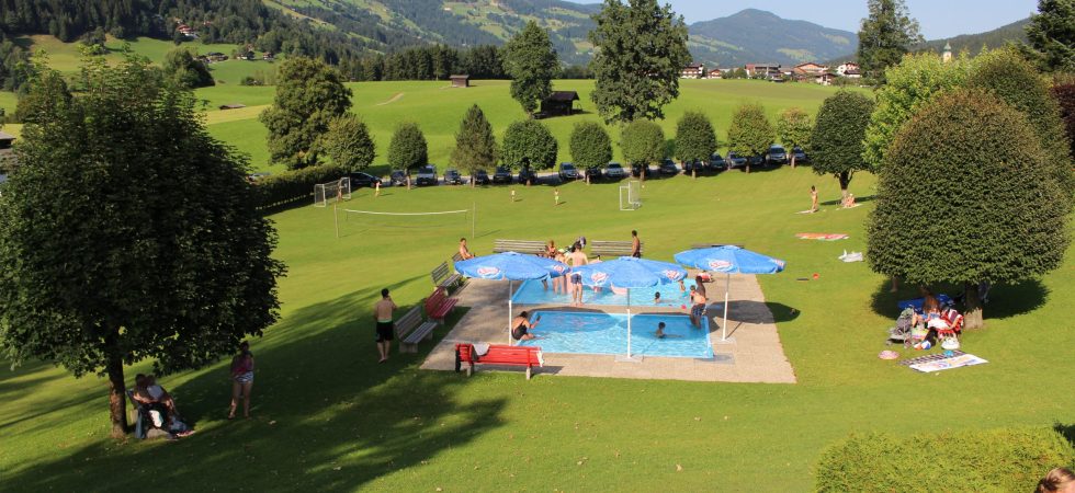 En naturskøn park med en lille pool, blå parasoller og mennesker, der slapper af, fanger essensen af en Sommerferie i Westendorf. Det græsklædte område er oversået med træer mod bølgende bakker og en fodboldbane, mens besøgende slapper af på græsset og nyder den solrige dag.