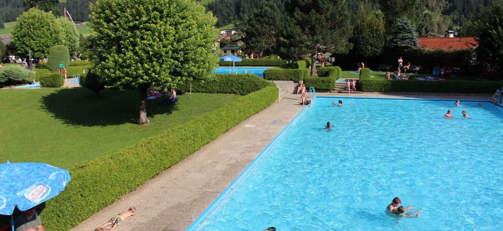 Under en solrig sommerferie i Westendorf er en udendørs swimmingpool omgivet af grønne hække og træer, hvor folk svømmer og soler sig. I baggrunden rejser sig frodige grønne bakker bag rekreative paraplyer.