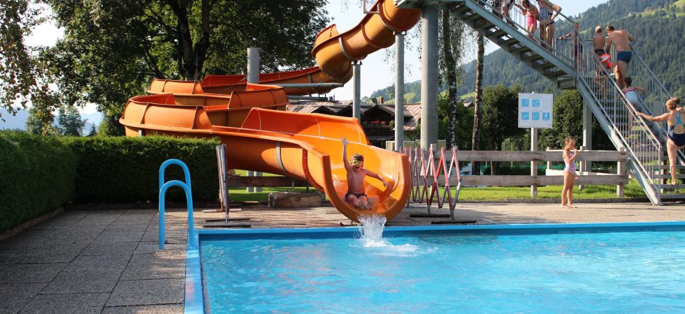 Midt i en solskinsdag perfekt til sommerferie i Westendorf glider en person yndefuldt ned ad den orange vandrutsjebane ned i den indbydende klarblå pool. Ivrige børn venter på deres tur på toppen, mens den naturskønne kulisse af træer og bakker fuldender den pulserende scene.