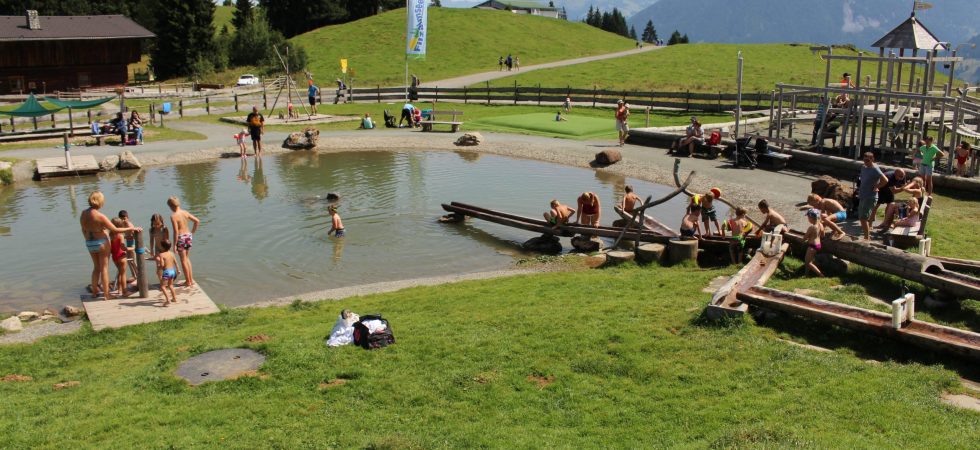 Folk nyder en solskinsdag i en lille park med en dam, der minder om Sommerferie i Westendorf. Børn leger i vandet og på trækonstruktioner, da voksne holder opsyn i nærheden. I baggrunden strækker frodige grønne bakker, spredte træer og en fjern bygning sig under en klar blå himmel.