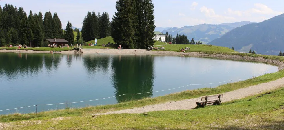 Et roligt alpine landskab med en fredfyldt sø omgivet af græsklædte bakker og fyrretræer. I det fjerne nyder folk deres sommerferie i Westendorf nær søen, med bjerge synlige under en klar blå himmel. En træbænk har udsigt over scenen.