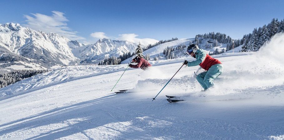 To skiløbere i farverigt gear suser ned ad en sneklædt bjergskråning i Maria Alm, med forrevne, snedækkede tinder og stedsegrønne træer under en klar blå himmel, hvilket giver den perfekte skiferie.
