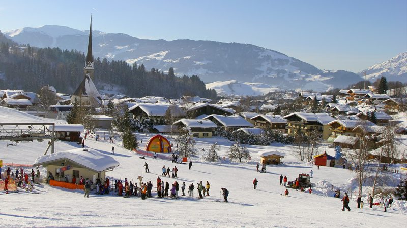 En malerisk sneklædt landsby med skiløbere på en skråning i forgrunden danner scenen for en skiferie i Maria Alm. En kirke med et højt spir titter frem blandt snedækkede huse og træer, mens majestætiske bjerge rejser sig under en klar himmel i baggrunden.