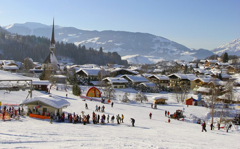 En malerisk sneklædt landsby med skiløbere på en skråning i forgrunden danner scenen for en skiferie i Maria Alm. En kirke med et højt spir titter frem blandt snedækkede huse og træer, mens majestætiske bjerge rejser sig under en klar himmel i baggrunden.