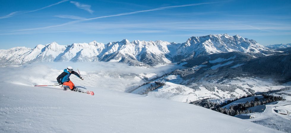 En skiløber i pulserende gear går ned ad en sneklædt skråning med snedækkede bjerge i baggrunden under en klar blå himmel, og fanger spændingen ved at stå på ski under en skiferie i Saalbachs fantastiske alpine landskab.