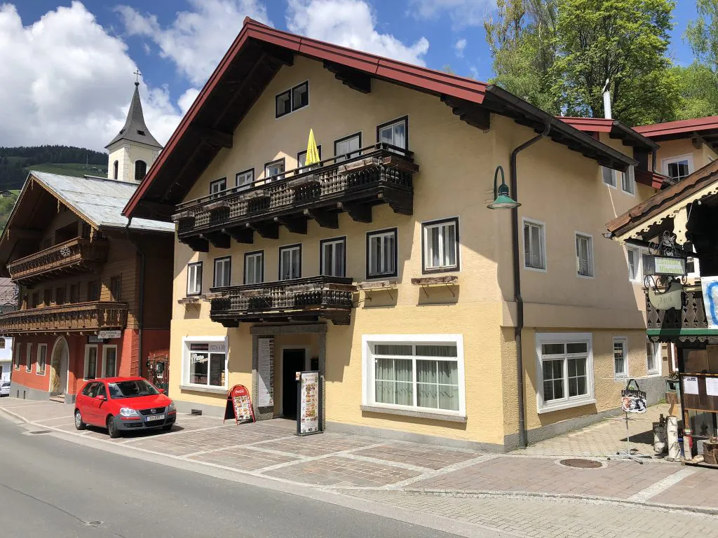Et malerisk gadebillede i Saalbach med en traditionel alpebygning med træbalkoner og en beige facade. En rød bil holder parkeret foran, og et kirketårn rejser sig i baggrunden. Den delvist overskyede himmel sætter gang i det frodige grønne område og fanger charmen ved skirejser-stemning.