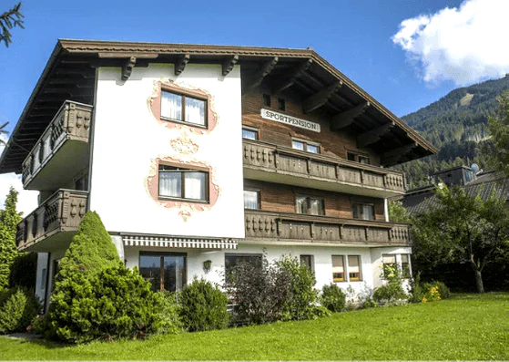 En traditionel alpehytte med tre etager, dens træbalkoner prydet med udsmykkede detaljer, tilbyder de perfekte rammer for en Sommerferie i Westendorf. Set mod frodige grønne områder og et majestætisk bjerg under en klar blå himmel, byder et skilt dig varmt velkommen til Sportpension.