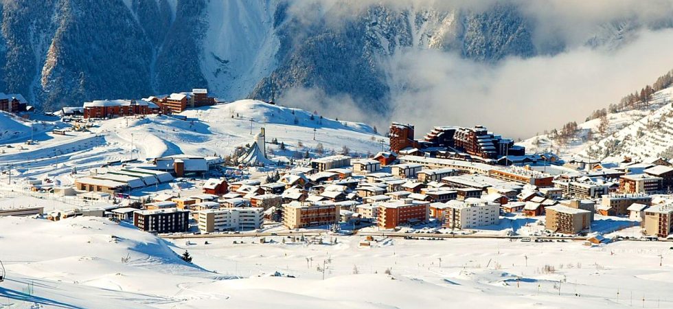 En sneklædt bjerglandsby beliggende i en dal, omgivet af maleriske tinder, tilbyder de perfekte rammer for en skiferie i Les Deux Alpes. Bygninger er spredt ud over landskabet med synlige skispor. Himlen er delvist overskyet, hvilket tilføjer kontrast til den lyse snedækkede scene.