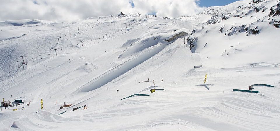 Et snedækket skisportssted i Les Deux Alpes har flere skilifte og snowboardere, der glider over ramper og skinner. Landskabet er dækket af uberørt hvid sne under en delvist overskyet himmel, mens forrevne bjerge står fremtrædende i baggrunden og tilbyder det perfekte skiferieflugtssted.