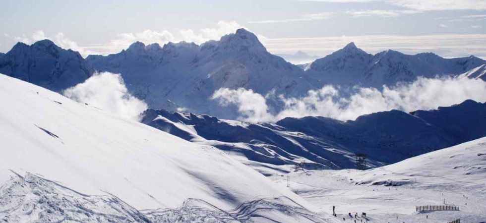 Den snedækkede bjergkæde under en klar himmel med spredte skyer indbyder til eventyr. Skiløbere på "skiferie i Les Deux Alpes" glider ned ad pisterne, mens fjerne tinder rejser sig majestætisk i baggrunden og skaber et fredfyldt vinterlandskab.
