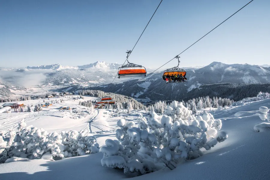 Et snelandskab i Westendorf viser fjerne bjerge under en klar blå himmel. To skilifter, fyldt med skirejse-entusiaster, stiger op over uberørte, snefyldte træer og pister. Nærliggende bygninger tilføjer farve og aktivitet til den rolige scene.