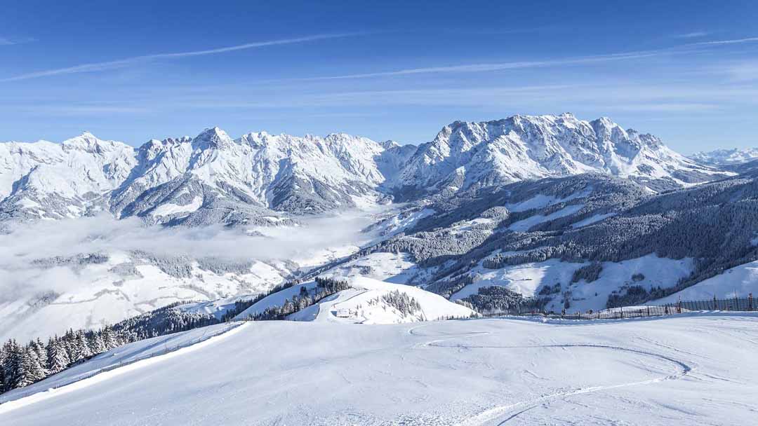 Et fantastisk vinterlandskab byder på snedækkede bjerge og dale under en klar blå himmel, perfekt til en skiferie i Maria Alm. Fyrretræer, der er støvet af sne langs skråningerne, og blide skyer svæver over toppene. Scenen er fredelig og uberørt.
