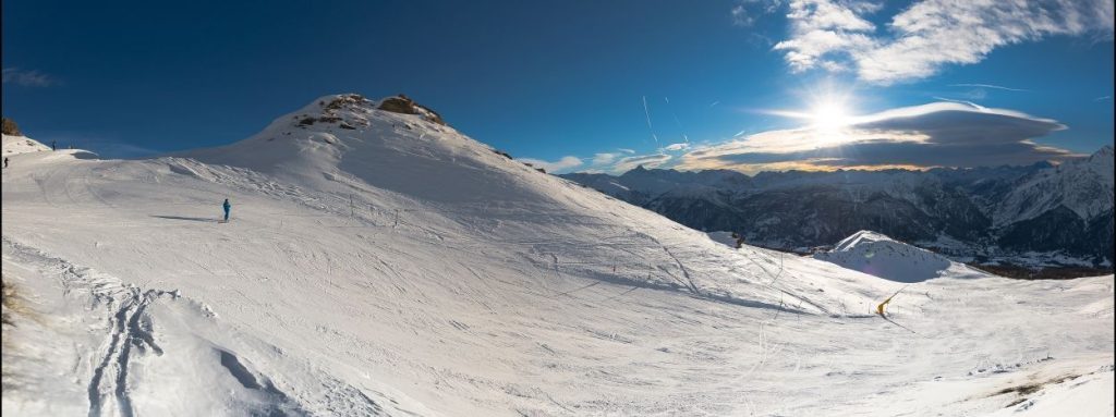 Panoramaudsigt over et snedækket bjerglandskab i Wagrain under en klar blå himmel. En ensom skiløber er på vej ned ad en svag skråning, mens solen går ned og kaster lange skygger. Fjerne tinder og spredte skyer tilføjer dybde til sceneriet, der minder om en alpin sommerferie i Østrig.