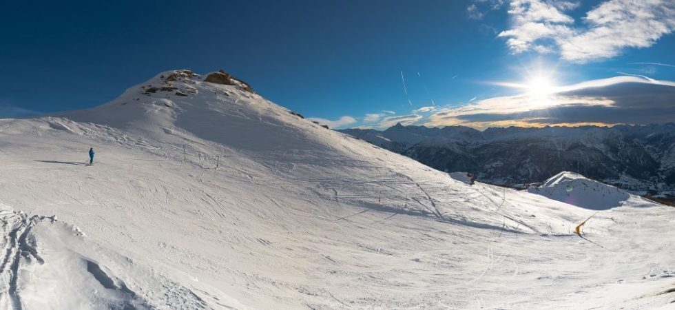 En panoramaudsigt over et snedækket bjerglandskab under en klar blå himmel. En enlig skiløber er synlig i det fjerne, måske nyder en skiferie i Westendorf. Solen står lavt på himlen og kaster lange skygger med bjerge, der strækker sig over horisonten.